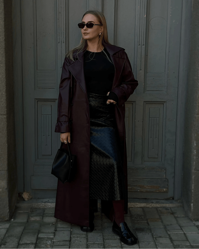Woman wearing a burgundy full-length faux leather trench coat open over a black knit and dark midi skirt